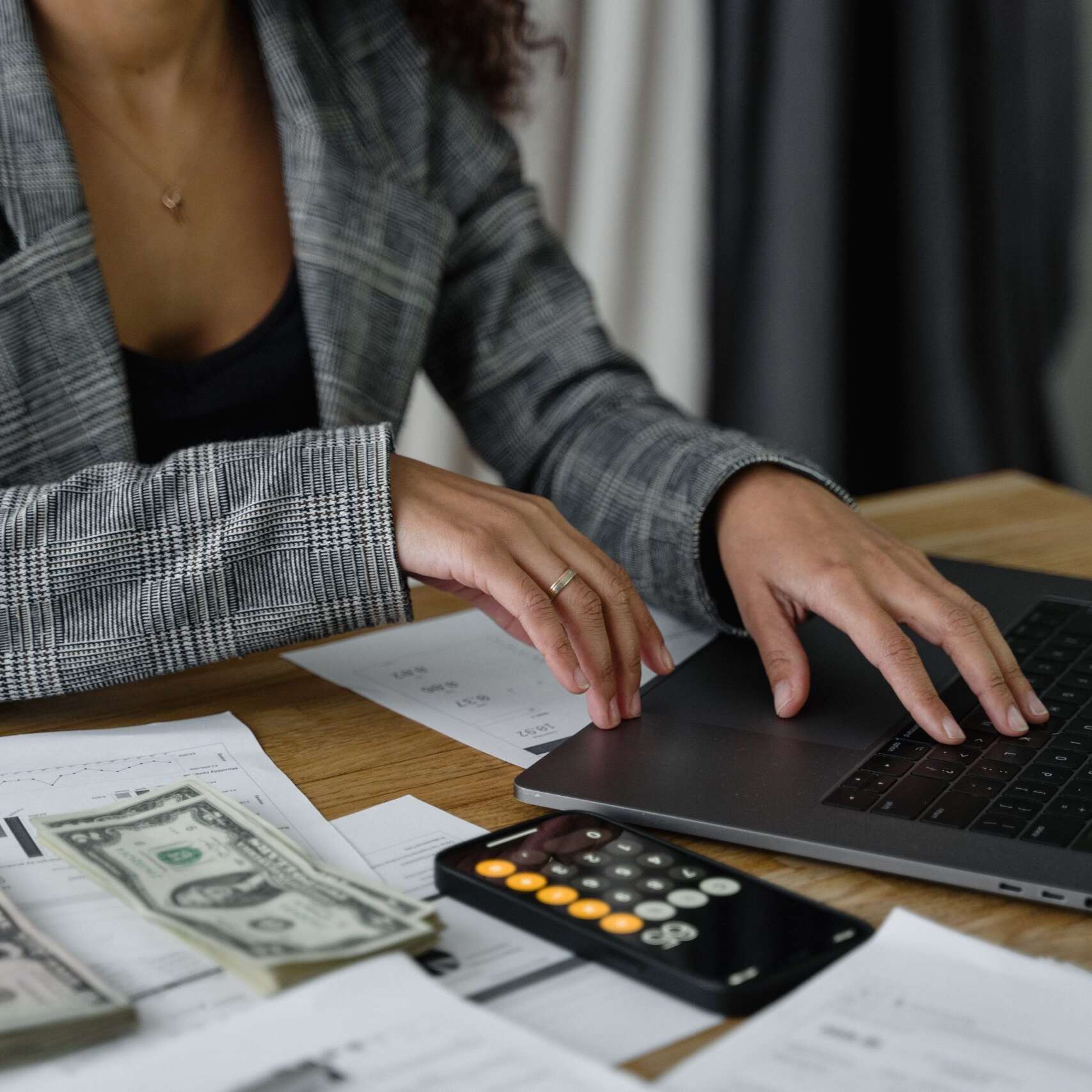 Woman typing on a laptop beside a calculator, financial papers and US dollar notes