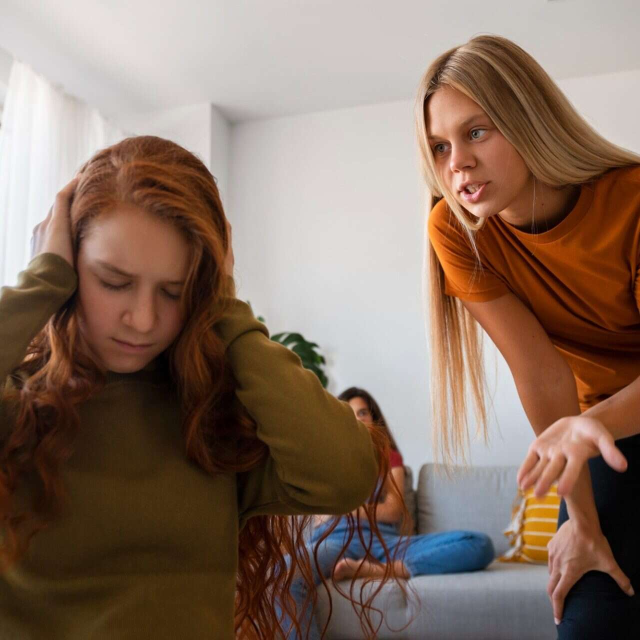 Young woman covering her ears while another woman leans over her and argues in a living room