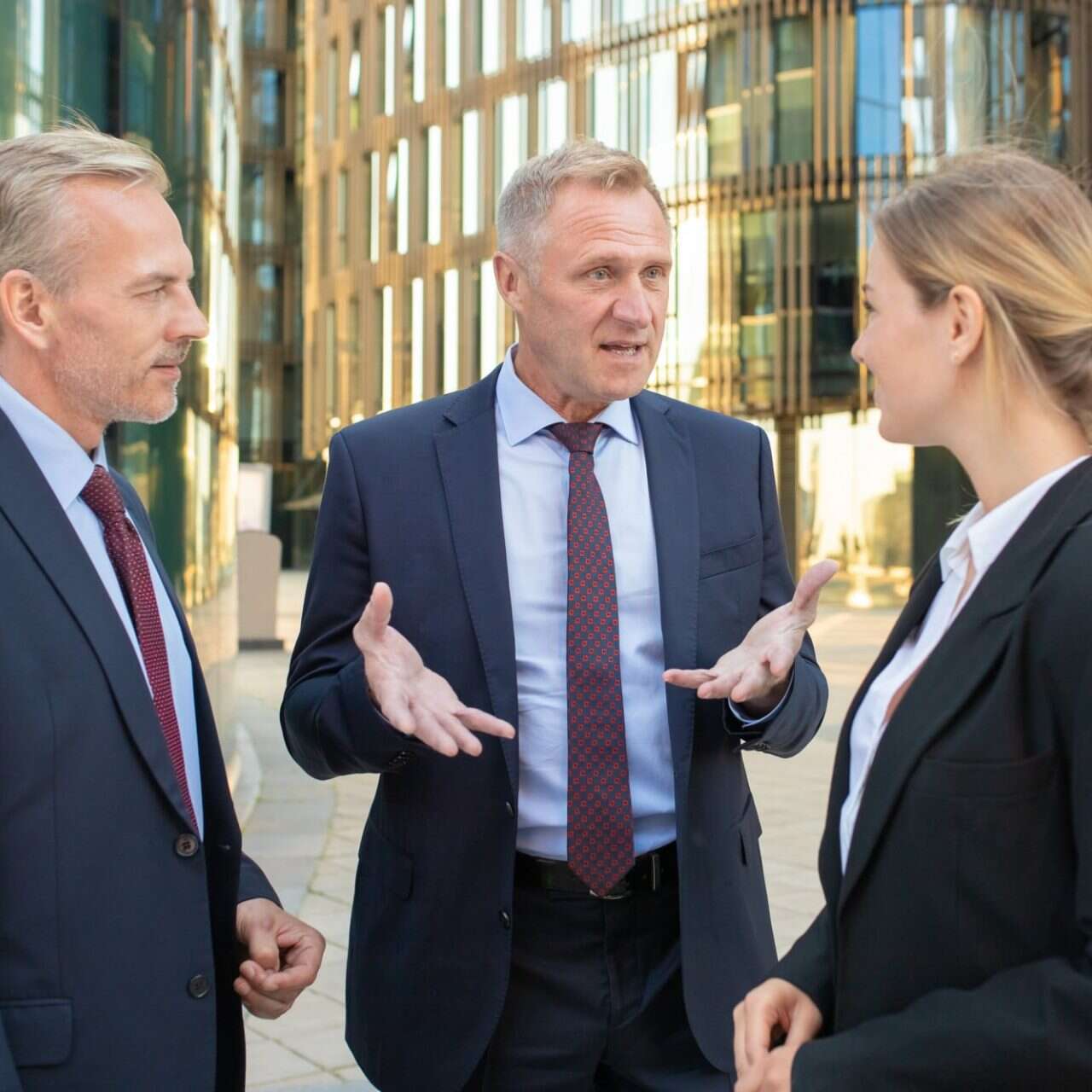 Two men and a woman in business suits talking outdoors in front of glass office buildings