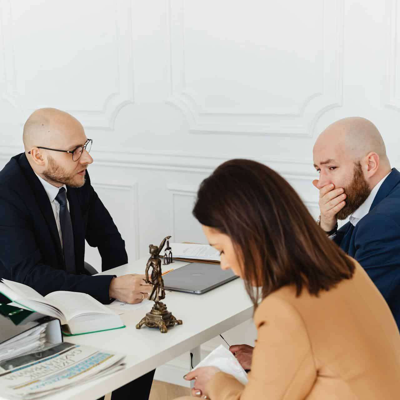 Lawyer speaking with a distressed couple at a white desk beside a small Lady Justice statue